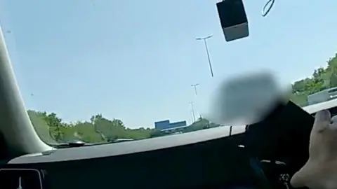 A hand holding a phone inside a car with a blurred out van in front on the road.