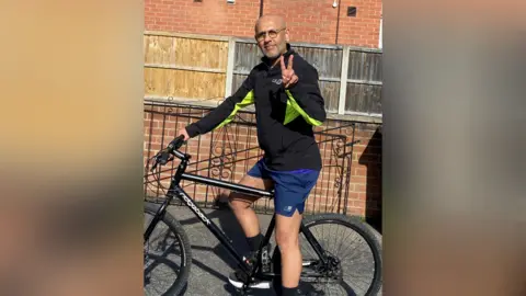 Supplied Shahid Akhtar in cycling gear on his bike wearing glasses and making a peace sign with his fingers