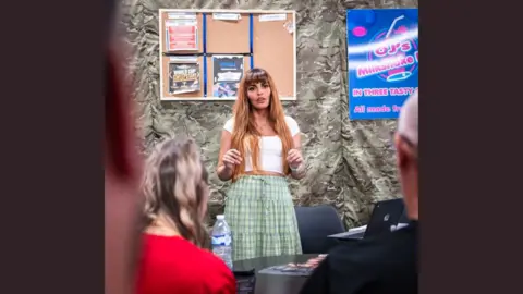 Slawek Wierny Picture of a young, long haired ginger woman in a white top and light green checked skirt. She is standing up and talking to a room. 