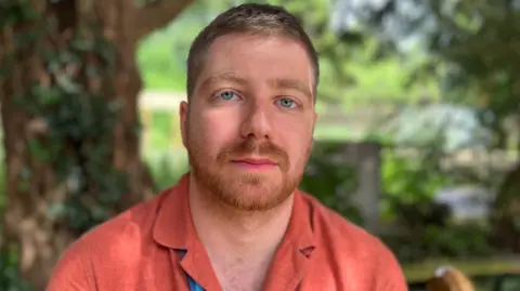 Headshot of Paddy Berryman, with a garden and tree in the background, he is wearing a coral coloured linen shirt