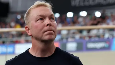 Sir Chris Hoy in a close-up image of his face, looking thoughtful and serious, his gaze off to the left. He is wearing a black T-shirt. A velodrome arena is blurred in the background.