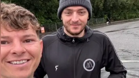 Grant Grego Will Fraser-Coombe is wearing a grey beanie hat, has short brown stubble and is wearing a gym-branded long-sleeved training jumper. Standing next to him is Grant Grego, who has light brown long hair and is wearing a grey hoodie. 