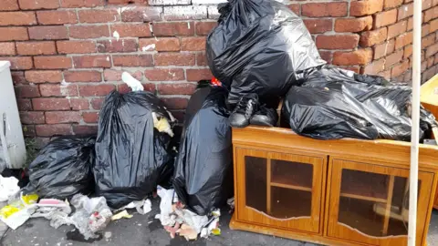 David Wall Piles of black bags and furniture have been left on a street corner in front of a red-block wall. 