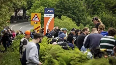 Fans line the side of the road at Hillberry on the the TT course. There is a road marker showing the section of the course as the road bends into the distance.