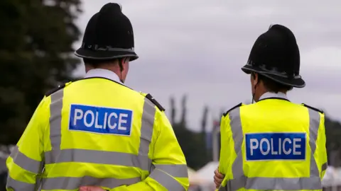 Two backs of two police officers can be seen. They are each wearing helmets and hi-vis jackets. It is a cloudy day.
