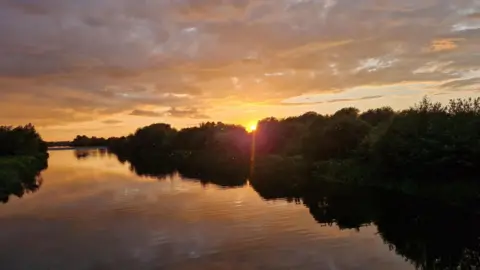 SunriseRunner WEDNESDAY - The sunsets over the river with trees on the bank as the river and sky glow orange