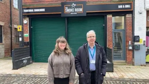 Sally Fairfax Beth Murrell - a woman with shoulder length blonde hair and a full fringe - is wearing glasses, a grey cardigan and smiling into the camera. Martin Taylor - standing next to her with short white hair - is wearing a black coat, blue lanyard and shirt, also with glasses on. They are standing on front of a building which says The Smoke Room in white lettering with a black background.