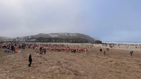 A large crowd of people wearing swimming trunks and costumes seen running across a beach towards the sea while people nearer the sea watch on.