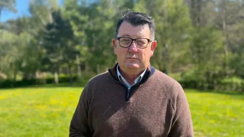 A man with glasses and dark hair wearing a brown zip up jumper