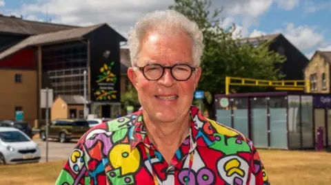 A man with white curly hair and round glasses wearing a colourful shirt