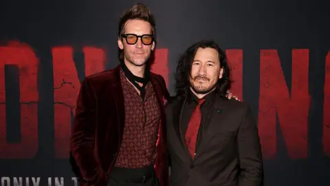 Getty Images. El actor Troy Baker y Mark "Markiplier" Fischbach asisten al estreno de "Iron Lung" en Los Ángeles. Están de pie frente a un fondo negro con letras rojas que dicen "Iron Lung". Markiplier viste un traje negro con corbata roja y luce una melena castaña hasta los hombros. Troy lleva una chaqueta de terciopelo color burdeos.
