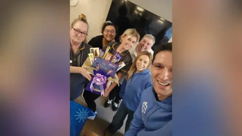 Albert Chait A group of people stand and smile together. Members of the pictures on the left hold a box of confectionary, given to them by those on the right of the image.