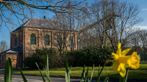 Sunderland City Council Washington's two-storey red brick F-Pit museum with a daffodil in the foreground. The metal headgear is still attached to the building, but is largely obscured by trees.