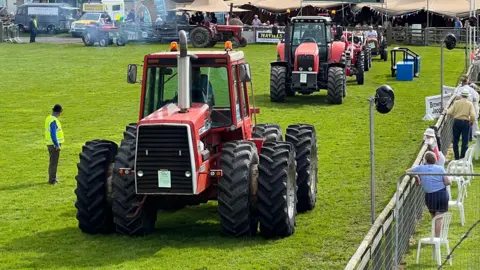 The image is taken at a previous Devon County Show, with tractors driving in a line, in one of the show rings. 