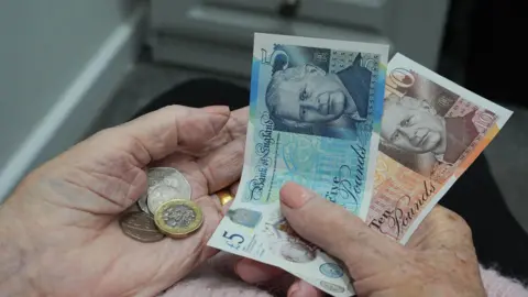 A five pound note, a ten pound note and coins are being held in the hands of an elderly person.