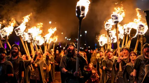 Ian Georgeson A man dressed as a viking holds a flaming torch above his head as he walks through a row of other men dressed in a similar way also carrying flaming torches above their heads.