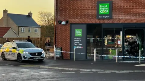 A lone police car parked outside a Co-op store. A police cordon tape is in place around the front of the store. The store is a red bricked building with a black entrance and some green signage. 
