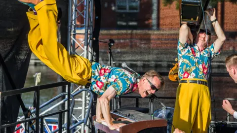 Stockton Council A man is jumping in the air as he plays the piano during a performance at Stockton Waterfront.
He is wearing yellow trousers, blue shirt with red and white flower print and sunglasses.