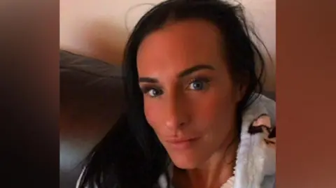 A selfie of Lisa Denton who has long dark hair and is slightly pouting into the camera. She is wearing a fluffy hoodie with dogs on them and sitting on a sofa.