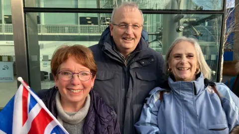Vikki Irwin/BBC Three people outside a glass-fronted building. The woman on the left holds a union jack flag and is wearing glasses and a purple coat. A man in the middle is wearing a dark blue jacket and a woman on the right is wearing a light blue jacket. They are all smiling at the camera.