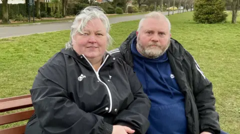 Two people sitting on a bench looking into the camera. One is a woman with short white hair, blowing over her face. She wears a black jacket with white piping. Next to her, a man with short blonde hair and a short beard wears a blue hoodie under a black jacket. Behind them is a grassy area with trees further back.