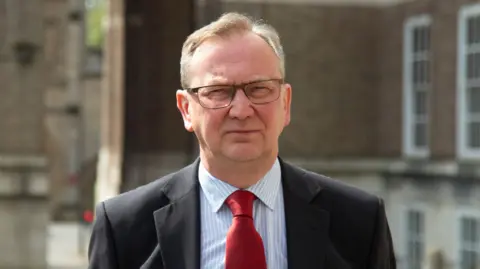 A head and shoulders shot of councillor Fabian Breckels, who is is squinting as he looks at the camera. He has short grey hair and wears rectangular, thin-rimmed glasses. He is wearing a black suit jacket, white and blue pin-striped shirt and a red tie. The council building is blurred behind him.
