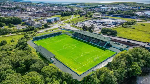 A CGI image of what the new Cumberland Sports Village stadium would look like from the air. The grass pitch has a large stand along the far touchline which runs roughly penalty area to penalty area. At the far end and along the near touchline are smaller stands. A road runs behind the large stand, with the rest of the stadium surrounded by trees and green space.