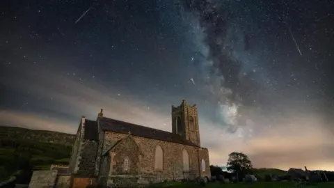 Patryk Sadowski A church is in the foreground with a dark sky above it. To the right of the picture is a Perseid meteor shower, which looks like fragments of white falling from the sky.