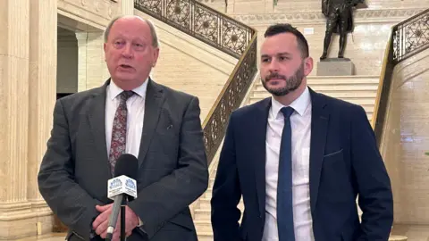 PA Media Jim Allister and Timothy Gaston standing in the Great Hall at Stormont. Jim is bald wearing a dark grey blazer, white shirt and burgundy floral tie. He is standing in front of a microphone. Timothy has short dark hair and beard, wearing a navy blazer, white shirt and navy tie. Behind them is a marble staircase with bronze statue at the top.
