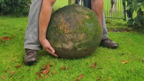 Calum Stott A person aims to pick up a round large boulder which is covered in moss. They are standing on a grassy field which has brown leaves on it.