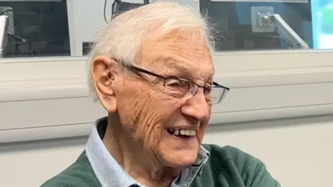 Noel Grugeon, a 99-year-old man with white hair and glasses, smiling with his teeth. He is sat in a radio studio, against a white wall and a window. He is wearing a green jumper.