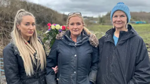 Three women in coats standing beside one another outside in front of a field looking at the camera. The sky is grey, the field is muddy and there are flowers behind them. 