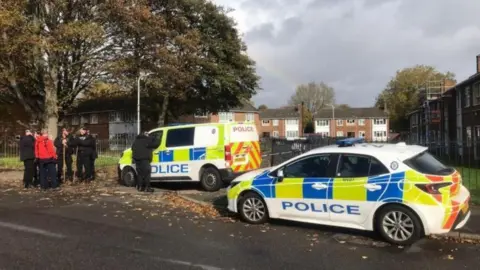 A police car and a police van are on a street. Police officers wearing all black, some holding long sticks, are on the left by a person wearing a red top. They are underneath a tree. Terraced homes can be seen in the background.

