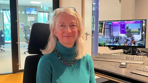 Sue Dougan/BBC Miranda Fyfe stares at the camera. She wears a green top and a 3 row necklace. She is sitting in an office style chair with a computer to the left 
