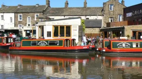 Two green and red Pennine Cruisers narrow boats on the Leeds and Liverpool Canal. They are moored beside waterfront bars with people sat outside.