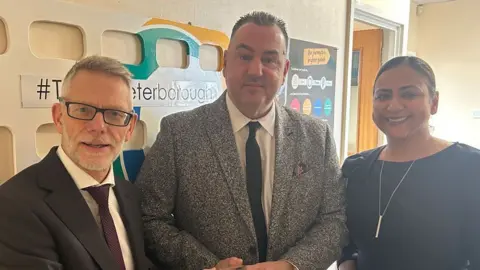 Stagecoach East Three people standing in a row looking towards the camera. On the left is Darren Roe in a dark suit, white shirt and maroon tie. He has a close-cropped  beard and is wearing rectangular dark framed glasses. Next to him is Paul McCarthy, in a black and white speckled jacket, white shirt and black tie. He is clean-shaven and has short dark spikey hair. Next to him is Hema Russell, who is wearing a dark dress or top and a long bar on a long necklace chain. She has pulled-back dark hair, tucked behind her ears.