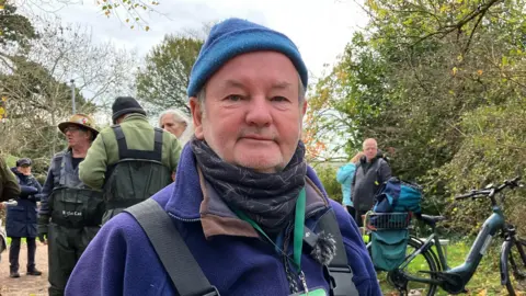 A man dressed in a blue beanie hat ,and dark blue fleece, stood in front of a group of people.