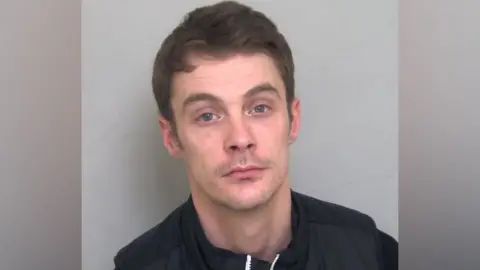 Cambs Police Charlie Martinson is looking at the camera in a police mug shot. He has short, slightly wavy brown hair, a little stubble and is wearing a dark top