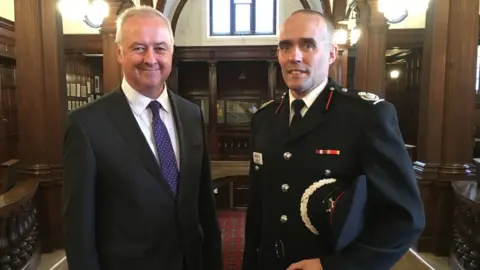 LDRS Two men, one in a dark suit and tie, the other in black formal firefighter uniform stand looking at the camera. The man in the firefighter uniform holds a formal cap under his arm.