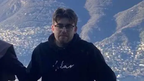 Sam Cartwright Sam Cartwright, wearing a black hoodie and glasses, with light brown hair. He is standing in front of rolling hills