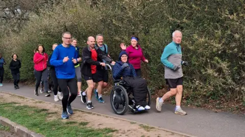 Amy Attwood A man in a powered wheelchair wearing a blue jacket and dark beanie is pushed along a paved path surrounded by a group of runners in colourful clothing. Bare hedgerow lines the path on the right and grass borders the left. Several runners are smiling and one woman waves at the camera.