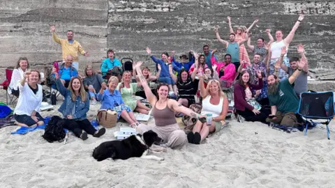 Kira Moisan Large group of people seated and standing on a sandy beach in front of a concrete wall, raising their arms toward the camera. A dog lies on the sand in the foreground, with picnic items and bags scattered around.