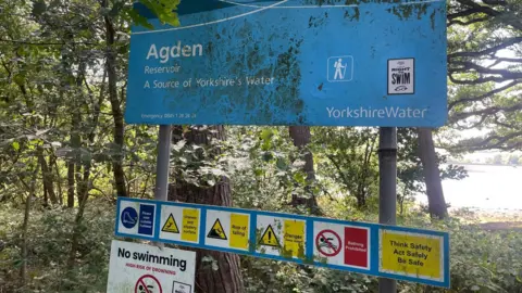 Simon Thake A large dirty blue sign stands amongst the trees with the sunshine and water poking through in the distance