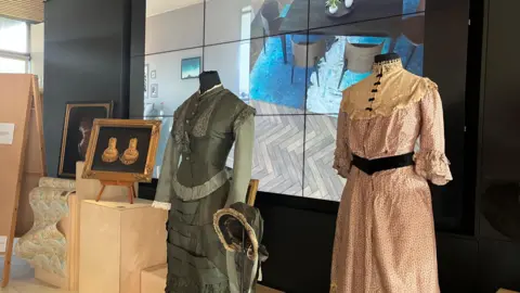 BBC Two of the dresses on stands from the Devonshire Dress Collection. There is a picture of jewellery next to the dark green dress on the left. There is also a screen behind the images.