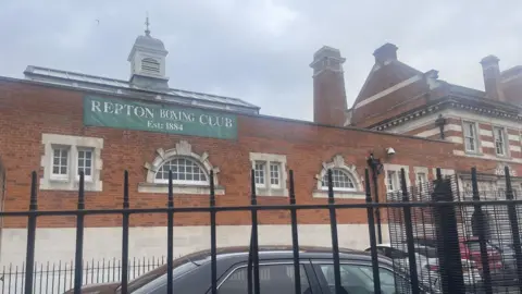 The front of a red and white brick building in east London. The building is behind black railings. There is a green sign with white writing above an arched window that reads Repton Boxing Club Est: 1884.