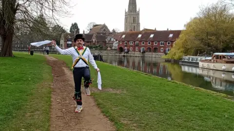 Duncan Bhaskaran Brown Duncan Bhaskaran Brown wearing Morris dancing attire, including a white shirt and black trousers, with bells tied around his shins. He has cross of green and yellow fabric over his shirt, a red tie around his neck, a black top hat and white handkerchiefs in each hand. He is dancing alongside the River Thames.