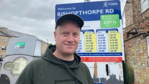 Josh, a Fulford resident using Bishopthorpe Road car park stands in front of the price information board and parking meter. He wears a cap and a green hoodie, with a black t-shirt underneath. 