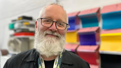 Tom Jackson/BBC Robert Whitehead is standing on the left. He has short grey hair and a long grey beard. He is wearing black framed glasses, a black T-shirt and jacket, and green lanyards. He is standing in front of a white wall which has different colour fabrics and folders.