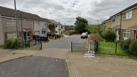 A google street view image of houses in a small close in Bradford. The houses are two-story and there is an open black metal gate across the top of the close. 