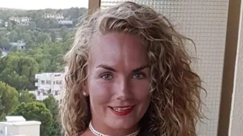 Family handout Carly on a balcony with trees and flat blocks behind her. She is smiling and has long thick curly blonde hair.